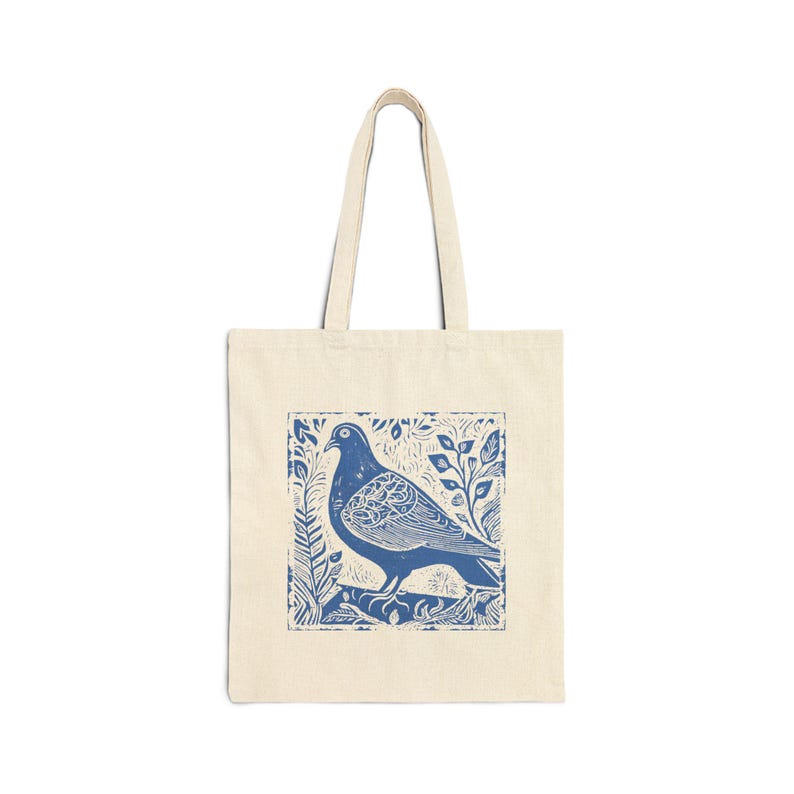 May include: Cream tote bag with a blue linocut print of a pigeon on a branch, framed by leaves. The bag has two handles and is made of a durable fabric. Ideal for carrying shopping or other everyday items.