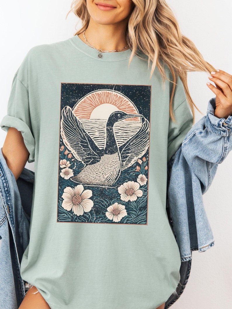 May include: A sage green t-shirt with a vintage-style graphic print of a bird in flight with a sunset and flowers.