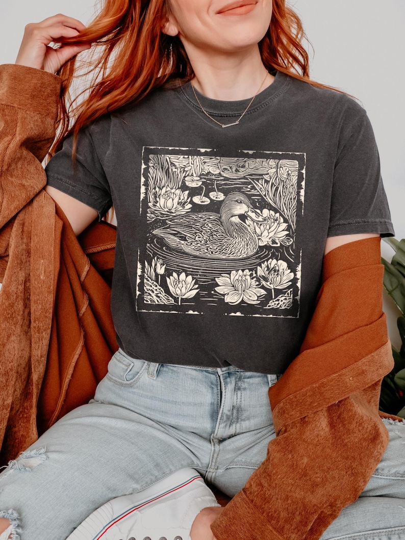May include: A dark grey t-shirt with a white print of a duck swimming in a pond with lily pads. The duck is surrounded by leaves and flowers.