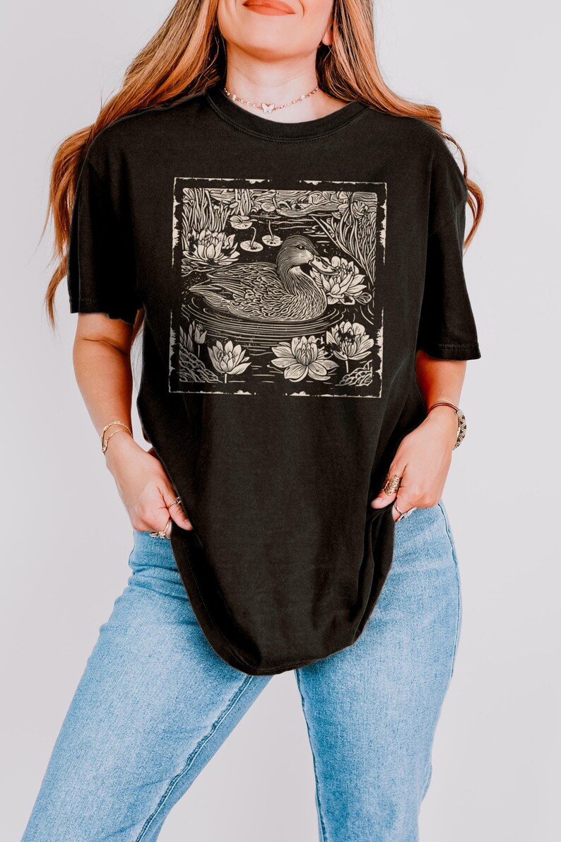 May include: A black t-shirt with a vintage-style illustration of a duck swimming in a pond with lily pads. The illustration is in a cream color and has a distressed look.