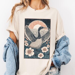 May include: A cream colored t-shirt with a vintage style graphic of a bird flying over a body of water with a sunset in the background. The graphic is surrounded by flowers.