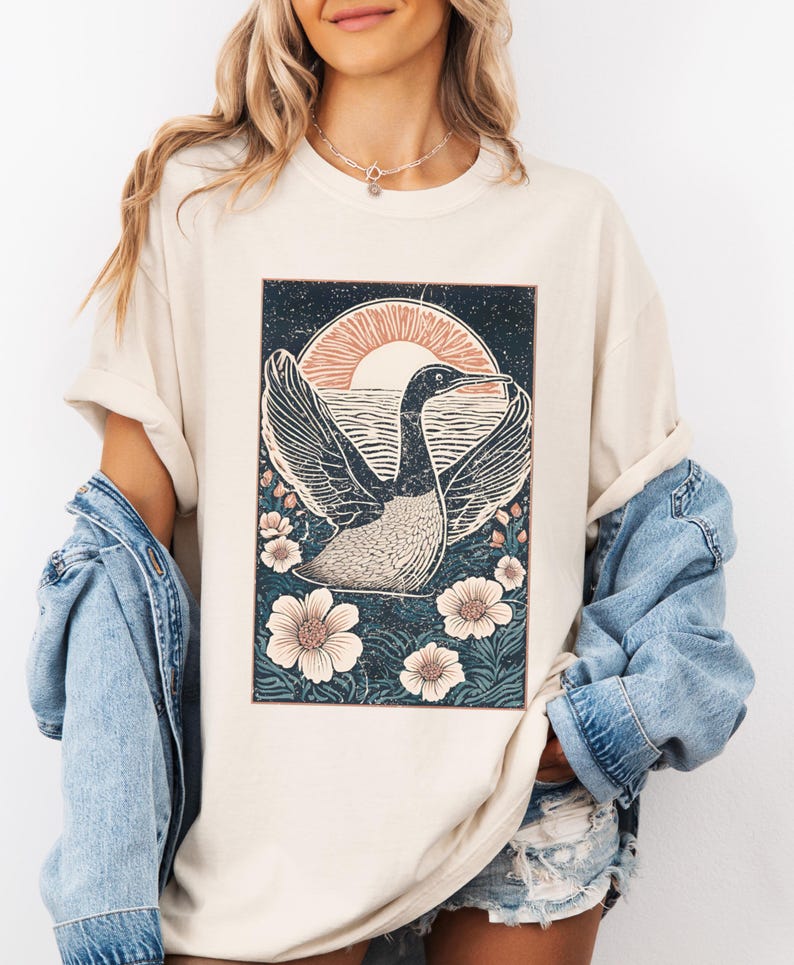 May include: A cream colored t-shirt with a vintage style graphic of a bird flying over a body of water with a sunset in the background. The graphic is surrounded by flowers.