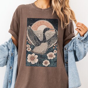 May include: A brown t-shirt with a graphic print of a bird flying over a body of water with a sunset in the background. The bird is surrounded by flowers.