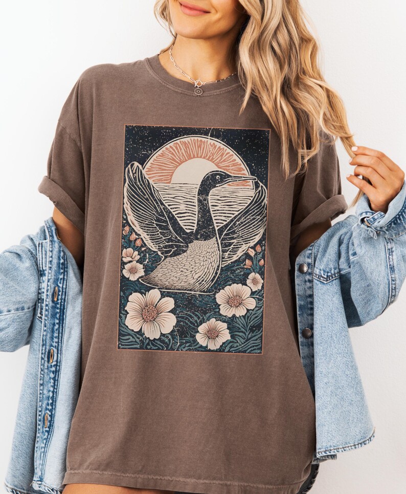 May include: A brown t-shirt with a graphic print of a bird flying over a body of water with a sunset in the background. The bird is surrounded by flowers.