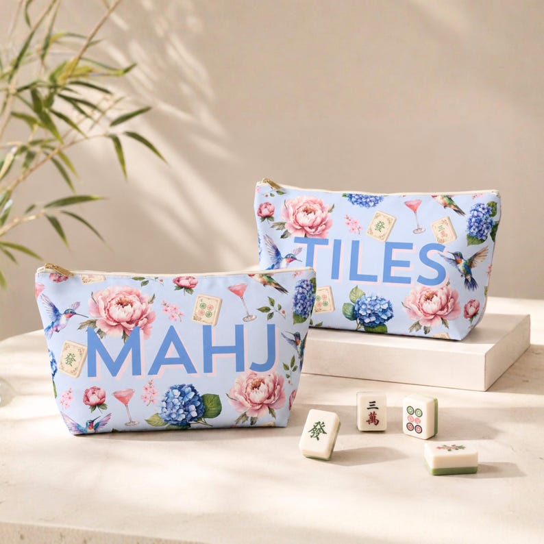 May include: Two light blue zippered pouches featuring a floral pattern with pink peonies, blue hydrangeas, hummingbirds, cocktail glasses, and mahjong tiles. One pouch displays the text MAHJ in blue, while the other displays the text TILES.