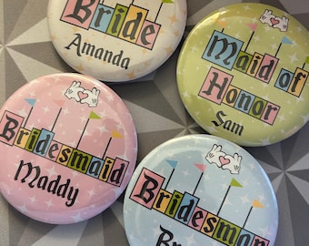 May include: Four round, colorful wedding party badges. The badges are light pink, light blue, light green, and off-white. Each badge has a different title: Bride, Bridesmaid, Maid of Honor, and Bridesman, with a name below each title. Each badge has a bunny graphic.