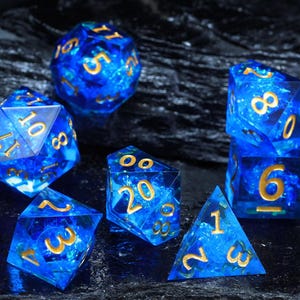 Op de afbeelding: Een set van zeven blauwe en gouden veelvlakkige dobbelstenen. De dobbelstenen zijn transparant met een wervelend blauw patroon en gouden cijfers. De set bevat een D4, D6, D8, D10, D12, D20 en een percentieldobbelsteen.
