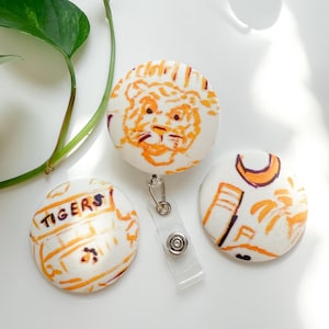 May include: Three white fabric button badges with orange illustrations. One badge features a tiger face, another has the word "TIGERS", and the third has a palm tree design. A clear plastic badge reel is attached to the tiger face badge.