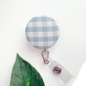 May include: A round badge reel with a light blue and white gingham pattern. The reel has a clear plastic clip and a metal swivel clip. A large green leaf is in the foreground.