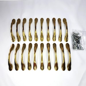 May include: A set of 18 gold-toned cabinet pulls with a decorative, ridged design. The pulls are arranged in rows on a white surface. A small bag of screws is visible to the right of the pulls. These pulls are likely for kitchen or bathroom cabinets.