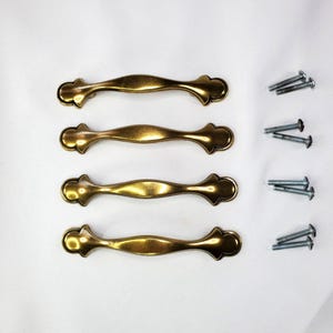 May include: Four brass-toned metal cabinet pulls with a curved, elongated shape and rounded ends, each featuring a smooth, polished finish. The set includes eight silver-colored metal mounting screws, with two screws provided for each individual handle.