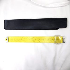 May include: A yellow slide rule with a clear plastic cursor and metal end brackets, accompanied by a black protective sleeve. The slide rule features printed numerical scales and markings across its length for mathematical calculations.