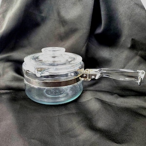 May include: A clear glass saucepan with a matching glass lid and a long, transparent glass handle. A wide, polished silver-tone metal band encircles the center of the pot, connecting the handle to the main body of the cookware vessel.