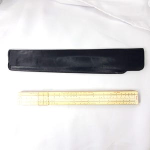 May include: A beige slide rule with a clear plastic cursor and printed numerical scales, accompanied by a black leather protective sleeve. The slide rule features various mathematical markings and text, including the manufacturer name.