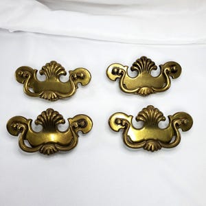 May include: Four antique brass drawer pulls with a decorative shell and scroll design. The pulls are a warm gold color and are arranged on a white background. These pulls would be suitable for a dresser or cabinet.