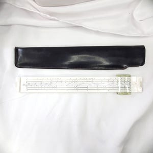 May include: A white slide rule featuring black numerical scales and a clear plastic cursor, accompanied by a long, rectangular black protective case. The slide rule displays multiple rows of precise markings for mathematical calculations.