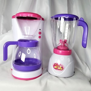 May include: A white toy coffee maker and blender set. The coffee maker has a purple carafe and accents, with pink and red details. The blender is white with a clear pitcher, purple lid and handle, and pink accents. The blender has the text "Just Like Moms".