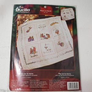 May include: A Bucilla Peace on Earth stamped lap quilt kit in a plastic package. The white fabric features a grid of nine squares with printed religious illustrations and text, measuring 45 inches by 45 inches. Includes instructions.
