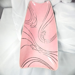 May include: A rectangular pink ceramic serving tray with concave sides and rounded corners. The surface features a black line art illustration of three stylized fish swimming amidst flowing, curved lines that span across the pink background.
