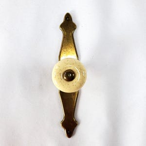 May include: A vintage-style cabinet pull with a cream-colored ceramic knob speckled with gray dots. The knob is set on a brass-toned metal backplate with decorative detailing. The overall design is symmetrical and classic.