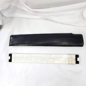 May include: A white slide rule with black numerical markings and a clear plastic cursor, accompanied by a long, slim black protective case. The slide rule features multiple scales and black end caps, providing a tool for manual calculations.