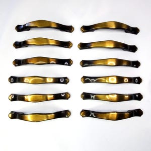 May include: Twelve antique-style cabinet handles with a curved design. The handles have a two-tone finish, with a gold center and dark brown edges. The handles are made of metal and are arranged in a grid pattern on a white surface.