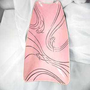 May include: A pink ceramic serving platter with a unique shape and black line art of fish swimming. The platter has a smooth, glossy finish and is ideal for serving appetizers or displaying decorative items.