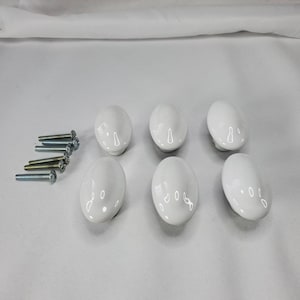 May include: Six white ceramic oval cabinet knobs with mounting screws. The knobs have a glossy finish and are arranged in a group. The screws are silver and gold-toned.