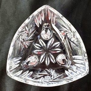 May include: A clear, triangular glass dish featuring intricate cut-glass patterns. The design includes a central floral motif surrounded by three circular indentations, with radiating starburst and leaf-like etchings along the beveled edges.
