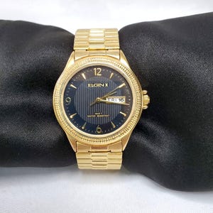 May include: A gold-toned Elgin watch with a dark blue face and gold hands. The watch displays the day and date, with the text "ELGIN" and "100FT WATER RESISTANT" visible. The watchband is also gold-toned.