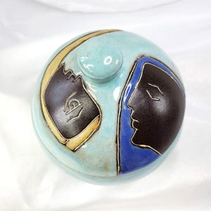 May include: A round ceramic lidded jar with a light blue glazed finish. The lid features two stylized dark brown human profiles facing each other, one outlined in gold and the other set against a deep blue background, with a central knob handle.