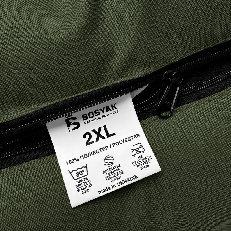 May include: Close-up of a green pet bed with a black zipper and a white tag. The tag reads "BOSYAK PREMIUM FOR PETS" and "2XL". It also includes washing instructions and "made in UKRAINE". The material is 100% polyester.