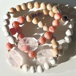 May include: Three bracelets stacked on top of each other. The top bracelet is made of white and grey beads. The middle bracelet is made of pink and red beads. The bottom bracelet is made of light brown wooden beads.
