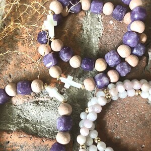 May include: Three bracelets with wooden beads and purple gemstone accents. Two bracelets have white cross charms. One bracelet has pink and white beads.