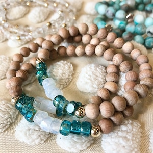 May include: Close-up of several beaded bracelets. The bracelets feature wooden beads, turquoise and clear glass beads, and gold-coloured metal accents. The bracelets are arranged on a textured, off-white surface.