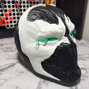 Może przedstawiać: Czarno-biała maska ​​o szczegółowym wzorze. Maska ma białą twarz i czarną głowę, z zielonymi akcentami wokół oczu. Prawdopodobnie element cosplay lub kostiumu.