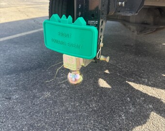 May include: A green plastic trailer hitch cover with the text "FRONT TOWARD ENEMY" in white lettering. The cover is attached to a black trailer hitch with a chrome ball.
