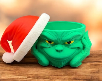 May include: A green Grinch-themed planter with yellow eyes and a hand resting on its cheek, paired with a red Santa hat with a white trim. The planter is on a wooden surface.