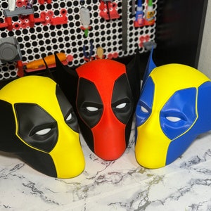 May include: Three superhero masks, one red and black, one yellow and black, and one blue and yellow. The masks are made of plastic and have a 3D design.