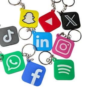 May include: A collection of colorful keychains featuring social media logos, including TikTok, Snapchat, YouTube, and Instagram. Each keychain has a silver keyring attached. The keychains are square or rectangular with rounded corners.