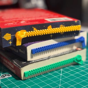 May include: Three books with colorful zipper-shaped bookmarks. The bookmarks are yellow, blue, and green, and are placed along the top edge of the books. The books have red, purple, and black covers.