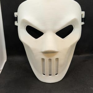 Può includere: Una maschera di plastica bianca con un design distintivo. Presenta ampie aperture angolari per gli occhi, un naso prominente e fessure verticali per la bocca. Cinghie nere sono attaccate ai lati e sulla parte superiore per fissare la maschera.