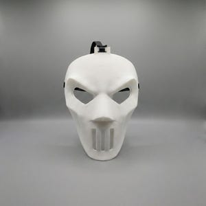 May include: A white, full-face mask featuring angular, aggressive eye cutouts and a prominent nose bridge. The lower portion of the mask includes a vertical, three-slotted grill design over the mouth area, with a black strap attached at the top.