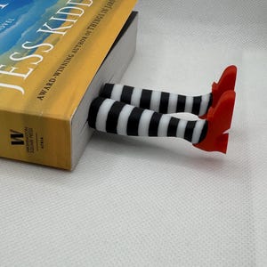 Peut inclure: Un accessoire de livre fantaisiste avec des jambes rayées noires et blanches et des chaussures rouges sortant d'un livre. Le livre a une couverture jaune et bleue avec le titre "JESS KIDD" et le texte "AWARD-WINNING AUTHOR OF THINGS IN JARS".