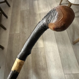 Könnte beinhalten: Ein handgefertigter Spazierstock mit einem dunklen, strukturierten Schaft und einem abgerundeten, braunen Kopf. Der Stock hat ein natürliches, gebogenes Design und ein beiges Band in der Nähe des Sockels. Die Gesamtlänge beträgt ca. 91 cm.