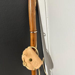 May include: A wooden walking stick with a silver ring and a small, light brown leather pouch attached. The pouch has a button closure and a logo. The stick is leaning against a dark wall.