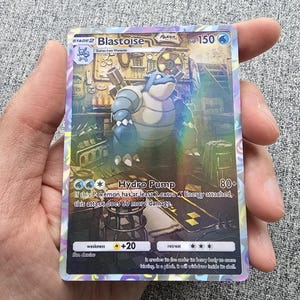 Peut inclure: Une carte Pokémon Blastoise bleue et grise du Jeu de Cartes à Collectionner Pokémon. La carte présente une illustration de Blastoise dans un décor steampunk. Le texte de la carte indique "Étage 2 Blastoise, Évolue de Tortank, PV 150, Hydrocanon, 80+, Si ce Pokémon a au moins 2 Énergies Eau supplémentaires attachées, cette attaque inflige 60 dégâts supplémentaires."