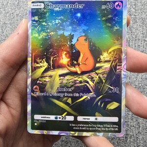 May include: A holographic Pokemon trading card featuring Charmander, a fire-type Pokemon. The card has a blue, purple, and pink background with a starry night sky. Charmander is sitting on a grassy patch with a small flame coming from its tail. The card text reads "BASIC Charmander HP 60 Ember Discard a Energy from this Pokemon. 30".
