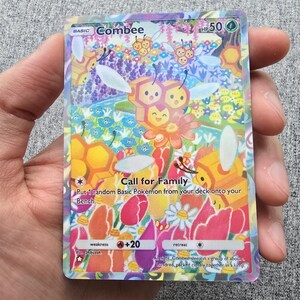 May include: A Combee Pokémon trading card with a colorful illustration of bees, flowers, and honeycomb. The card features the text "Combee", "Call for Family", and other game-related details. The card is held in a hand.