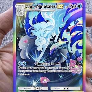 May include: A holographic Alolan Ninetales EX Pokémon card. The card features a blue and white fox-like creature with a rainbow border. The card text includes "Binding Snow" and "HP 150".